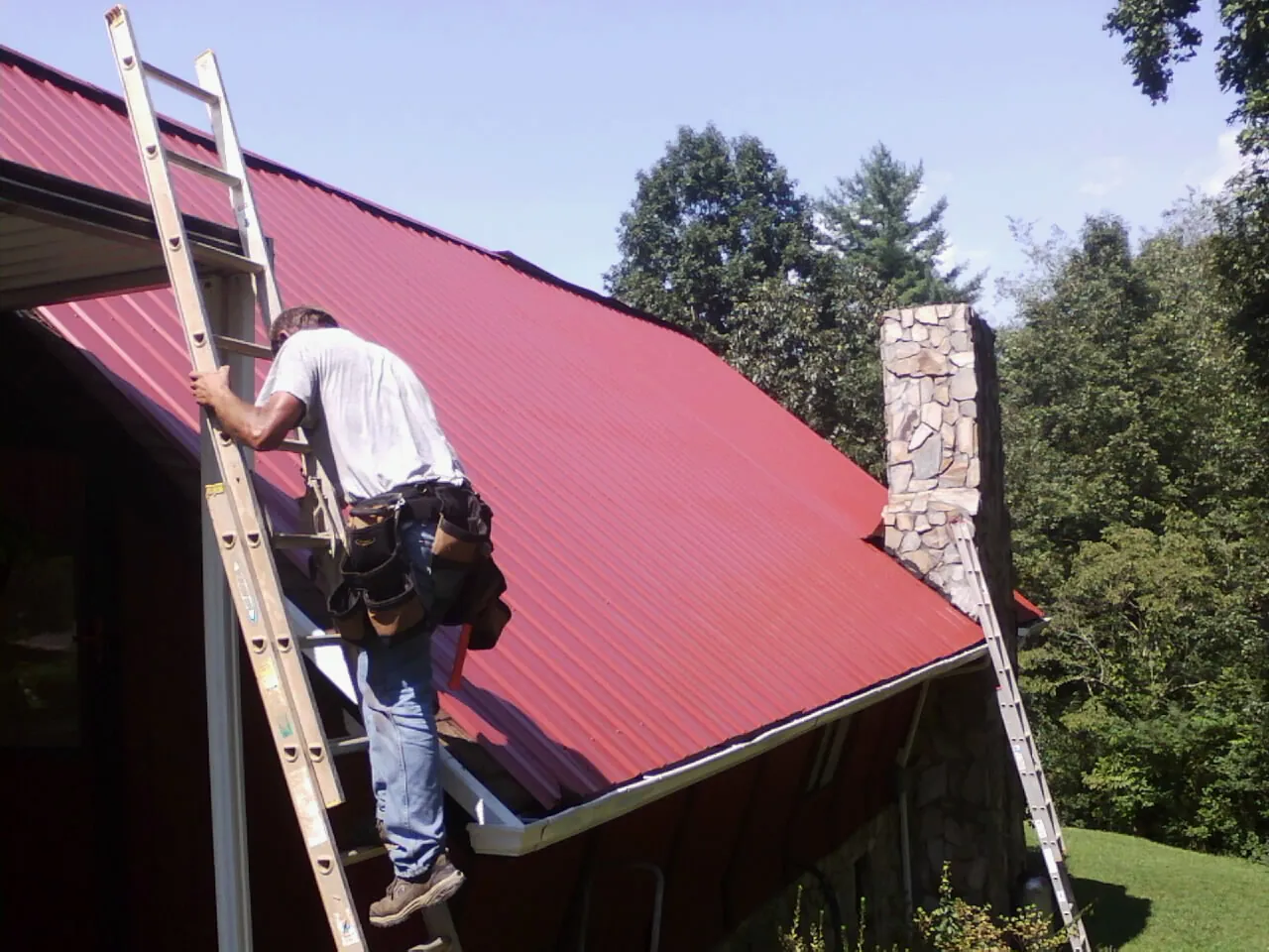 Roofer on a ladder beside a red metal roof and stone chimney — Soffit & Fascia Repair by Roofing Brothers in Central Islip